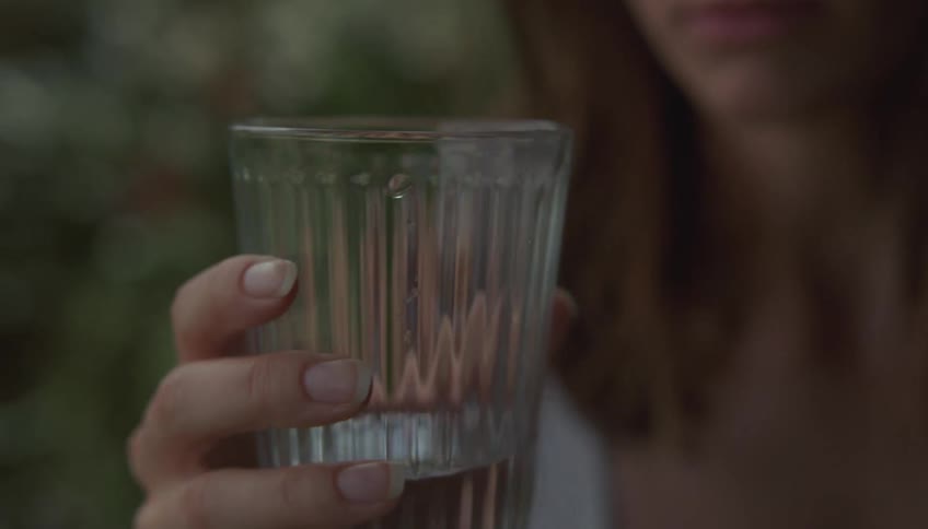 A woman takes a slow sip from a ribbed glass of water while standing outdoors in soft natural lightafter drinking she lowers the glass and begins to tilt it pouring the remaining liquid onto the ground