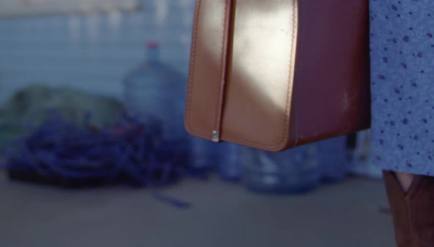 Woman walking with a travel suitcase beside a house wearing boots and a blue dress