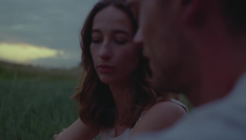 A woman with brown hair sits in a grassy field at dusk talking to a man and smoking a cigarette while the fading light of sunset glows on the horizon behind them