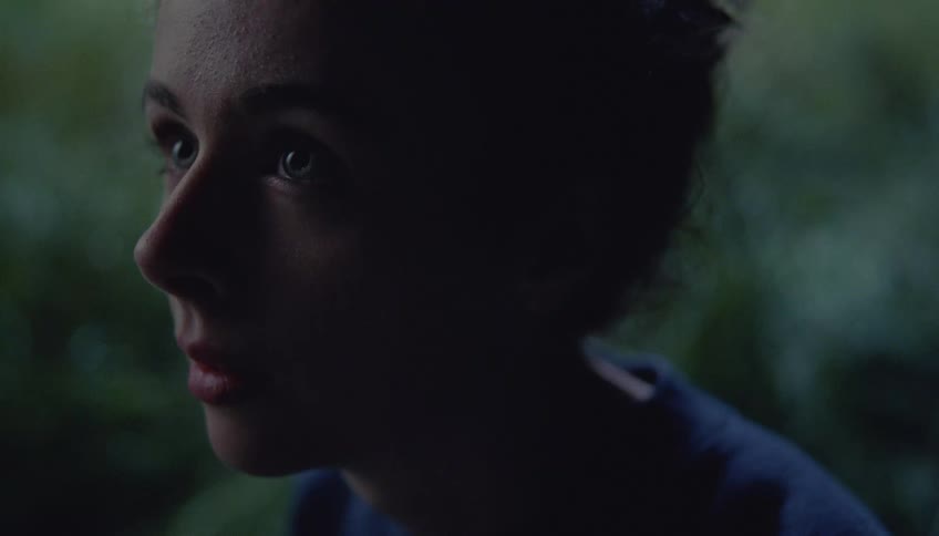 Videoa close view of a woman with dark hair pulled back looking off to the side with a serious expression in a dimly lit setting with green foliage in the background