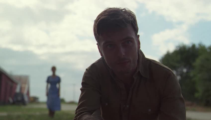 A man in a brown shirt crouches on the grass holding a small object while a woman in a blue dress stands in the distance near a farm building under a bright sky