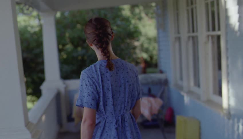 Woman exits the front door carrying a suitcase onto the porch on a summer day