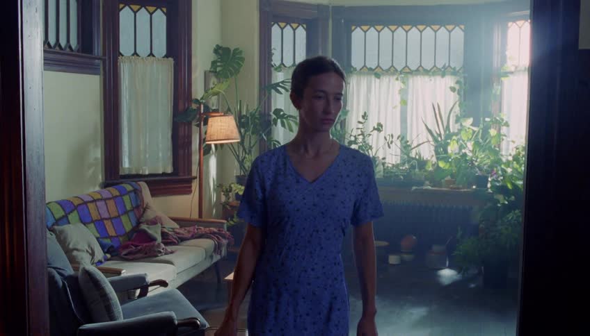 A woman walks out of a sunlit living room filled with houseplants carrying a suitcase