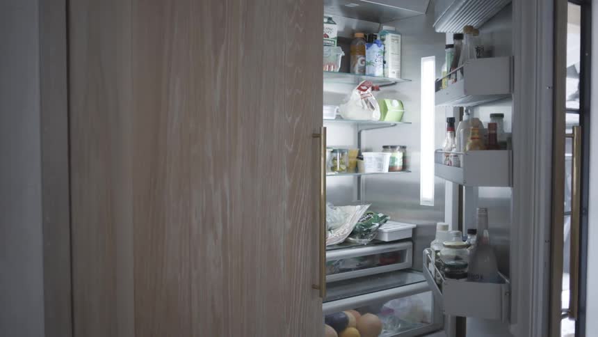 A large wooden cabinet door is opened to reveal a fully stocked refrigerator hidden behind the panelingthe door remains open for several seconds showing various food items and bottles before it is pushed closed again