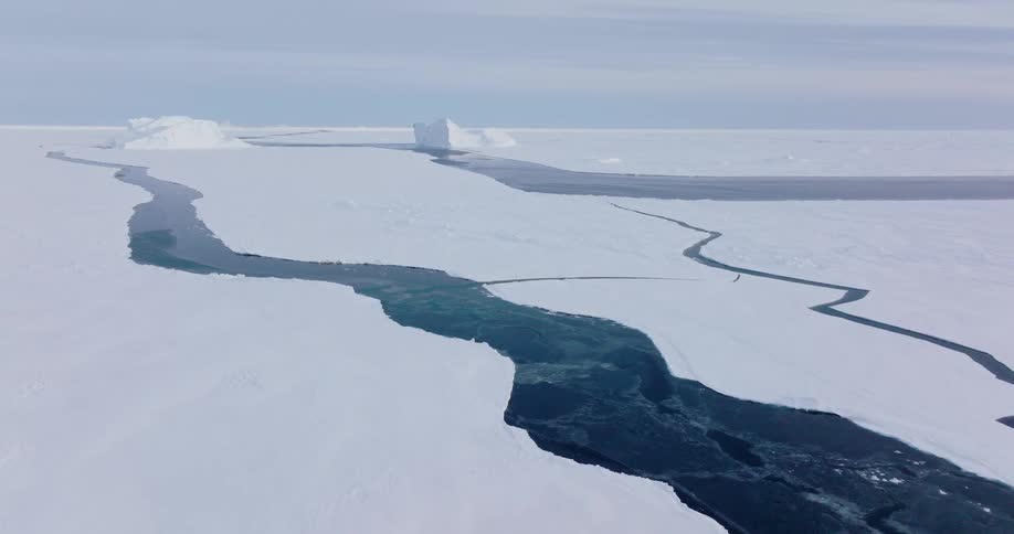 A vast sheet of arctic ice is fractured by winding cracks filled with dark blue water highlighting the stark contrast between solid ice and the open sea