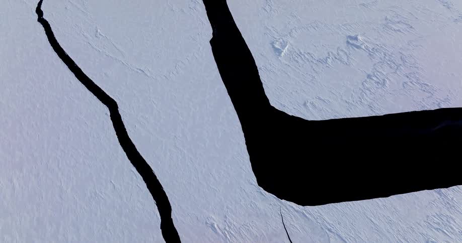 An aerial view shows two long dark cracks in a large field of white ice during the day