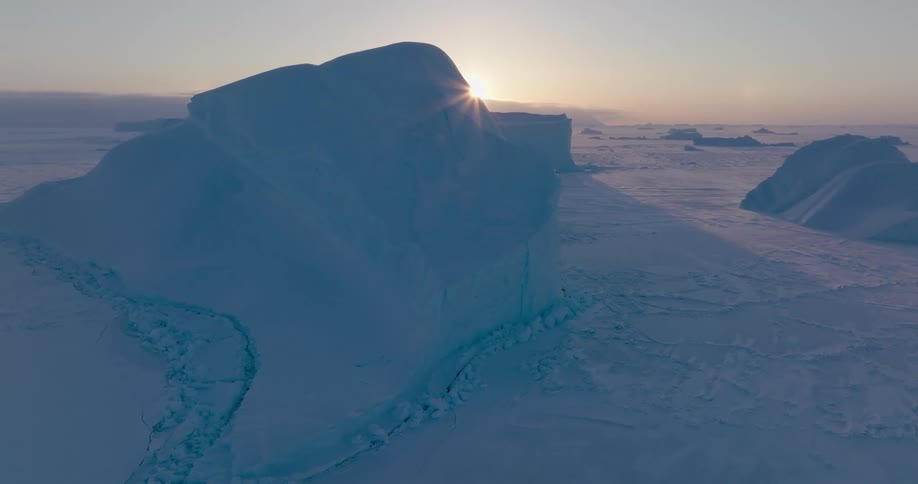 A massive iceberg rises from a frozen seascape bathed in soft blue light as the sun sets on the polar horizon