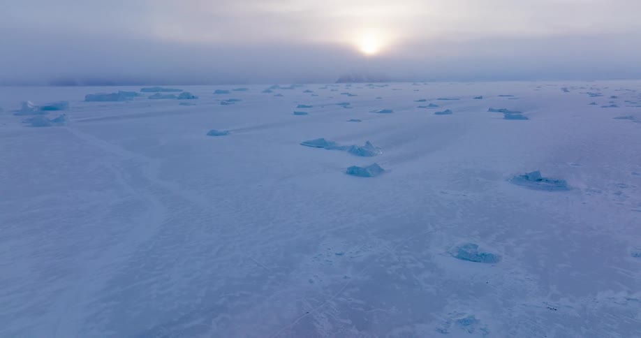 A wide view across a frozen landscape shows pieces of blue ice scattered on the snowy ground under a pale sun shining through a hazy sky