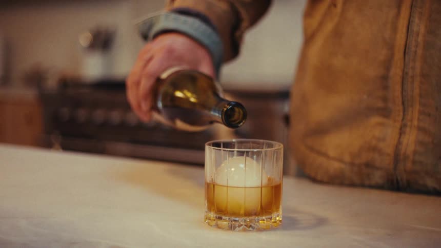 Hand pours an alcoholic drink from a bottle into a glass with ice on a countertop