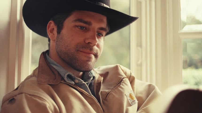 Man wears a hat and light jacket while looking at something offframe under natural indoor light