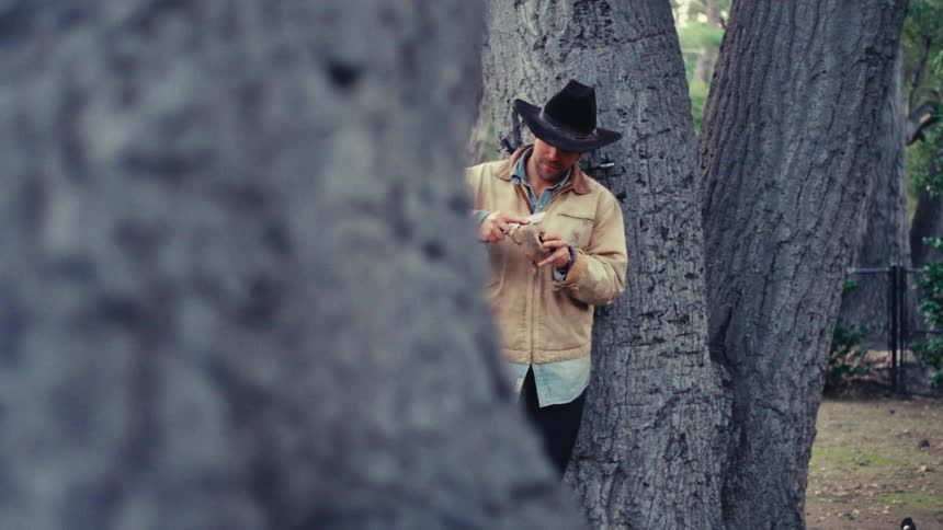 A man wearing a cowboy hat and a tan jacket stands partially obscured behind a large tree trunk he appears to be focused on something in his hands while leaning against the bark in a wooded area