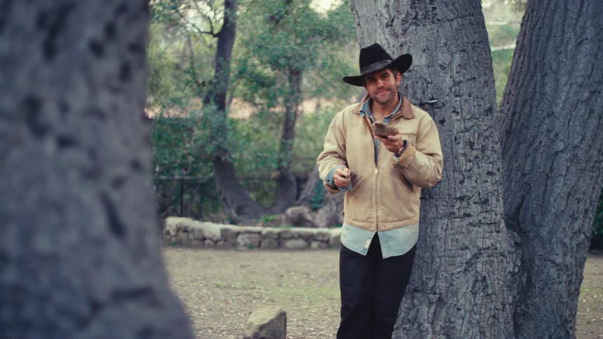 A man wearing a black cowboy hat and a tan jacket leans against a large tree trunk while holding a carved wooden object and smiling toward the camera in an outdoor setting