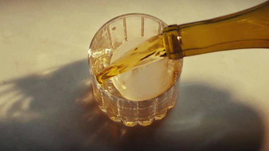 Amber liquid is poured from a bottle into a glass under direct warm lighting