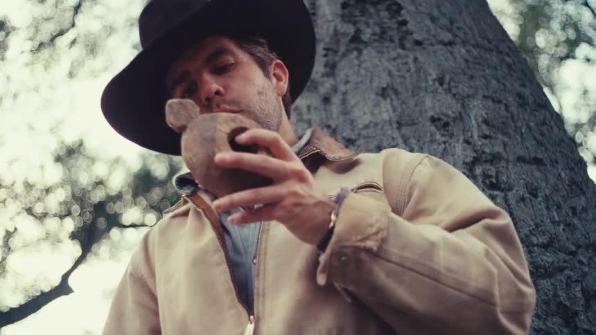 A carpenter wearing a black hat and tan jacket looks down while holding a carved wooden object in his hands outdoors near a large tree
