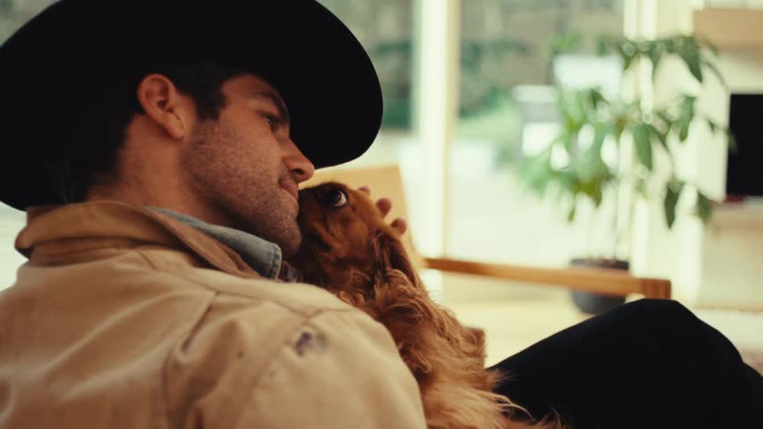 Man wearing a hat holds a dog on his lap in a welllit indoor setting