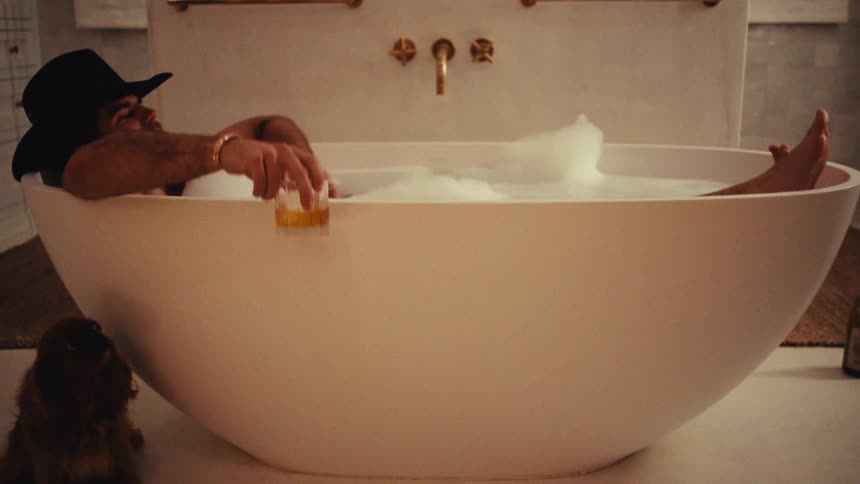 A man wearing a black cowboy hat relaxes in a white oval bathtub filled with bubbles while holding a glass of whiskey as a small dog sits on the floor nearby in a bright bathroom