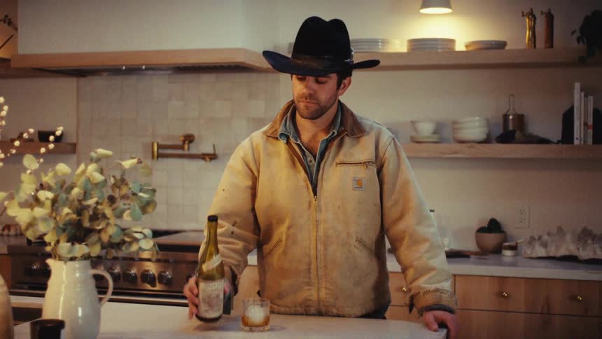 A man wearing a cowboy hat and tan jacket stands behind a kitchen island and pours a liquid from a glass bottle into a rocks glass with ice before looking directly into the camera lens while resting his hands on the counter surface
