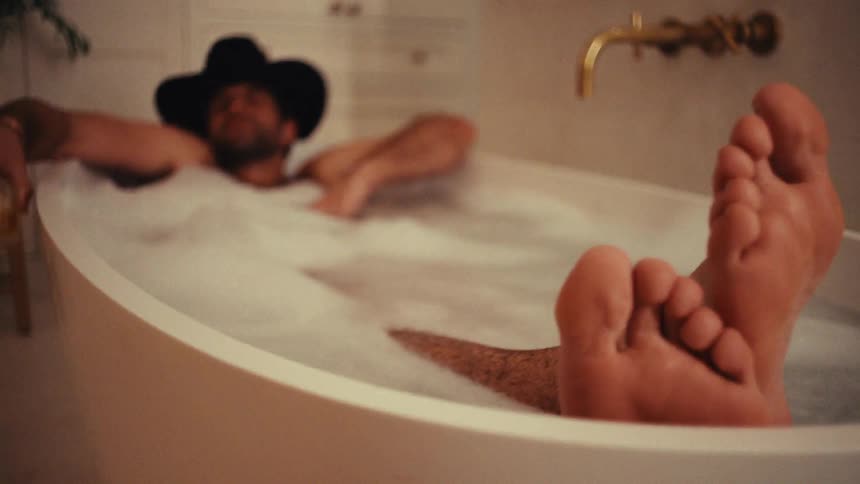 A man wearing a black cowboy hat relaxes in a white bathtub filled with bubbles and drinks from a glass of amber liquid with his bare feet resting on the edge of the tub