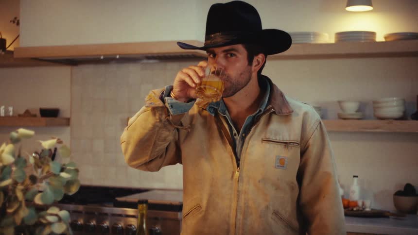 A view shows a man wearing a black cowboy hat and a tan jacket as he drinks from a glass in a kitchen during the day