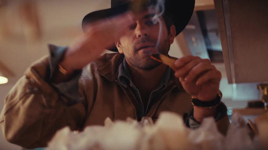 A man in a cowboy hat holds a burning piece of paper and calmly waves away the rising smoke with his other hand