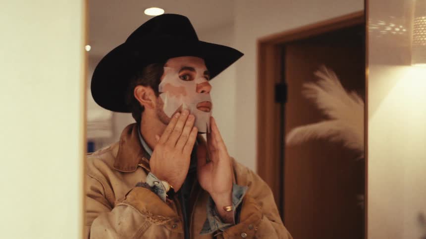 Man wearing a hat applies a facial mask in front of a mirror indoors