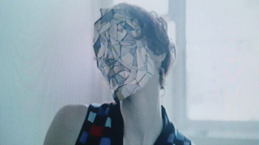 A person wearing a mask made of mirrored shards and a checkered vest moves their head in front of a bright window in a grainy video