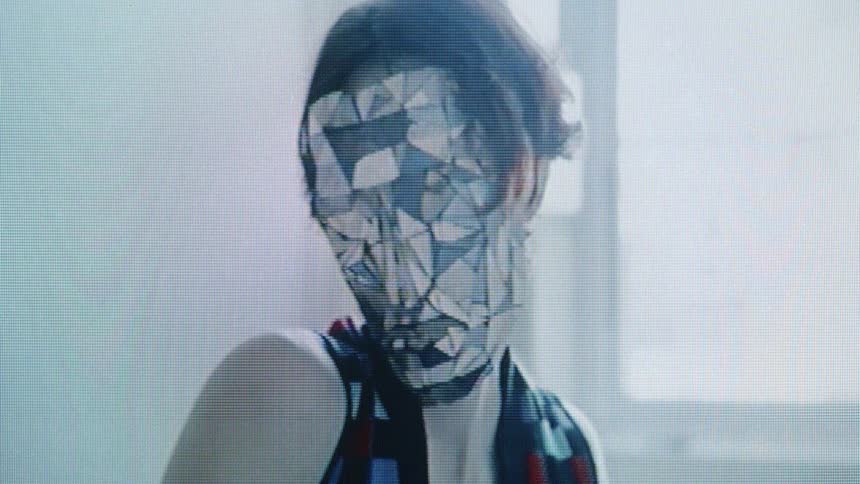 A person in a mirrored mask performs slow dramatic gestures in a softly lit interior