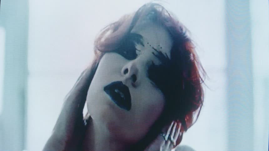 Woman with dark eye and lip makeup presses her hand against her head under strong backlighting
