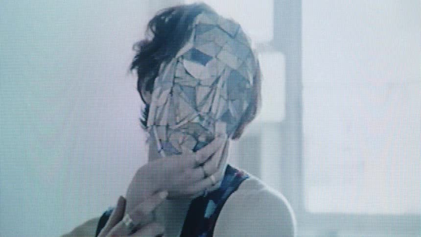 A view shows a person wearing a mask made of many small mirror pieces as they touch it while standing in a room during the day