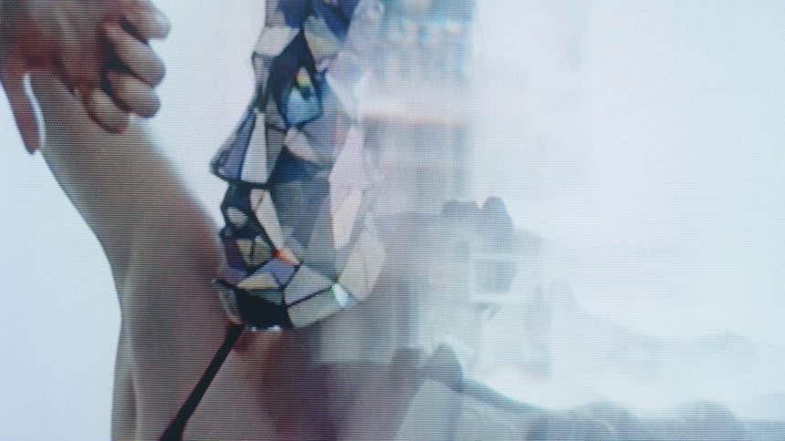 A mirror masked person performing slow eerie hand movements against a pale background