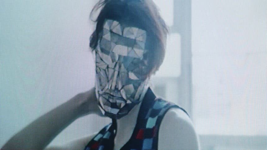 Adult man wearing a mirror mask and moving in a quirky way next to a window outdoors during day time