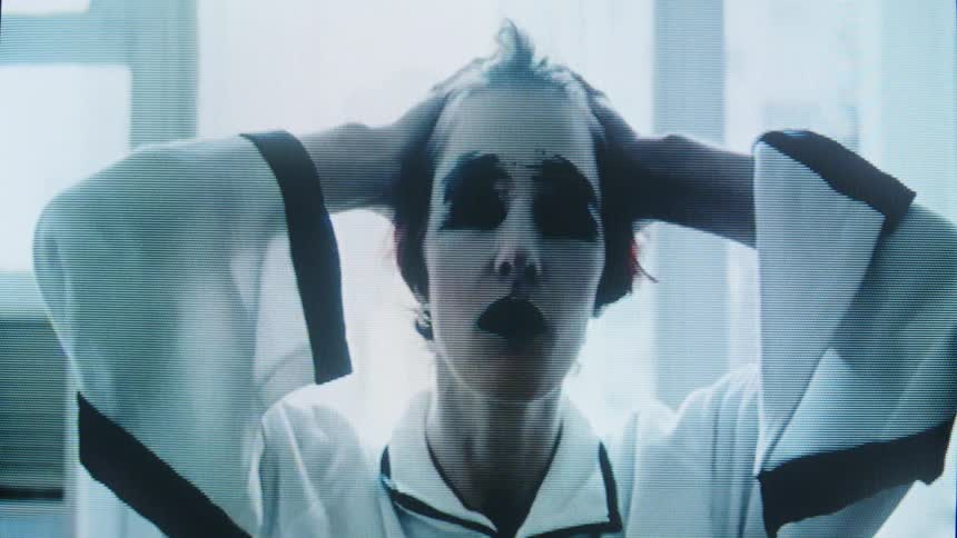Woman with dark facial makeup faces the frame in an indoor space lit by a window