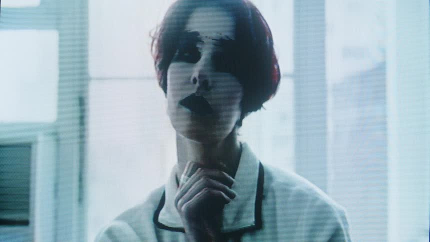 A person with dark face makeup performing slow surreal movements in soft lit room