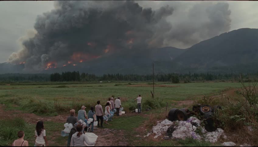 A line of people carrying supplies stands in a rural field as a massive wildfire burns nearby