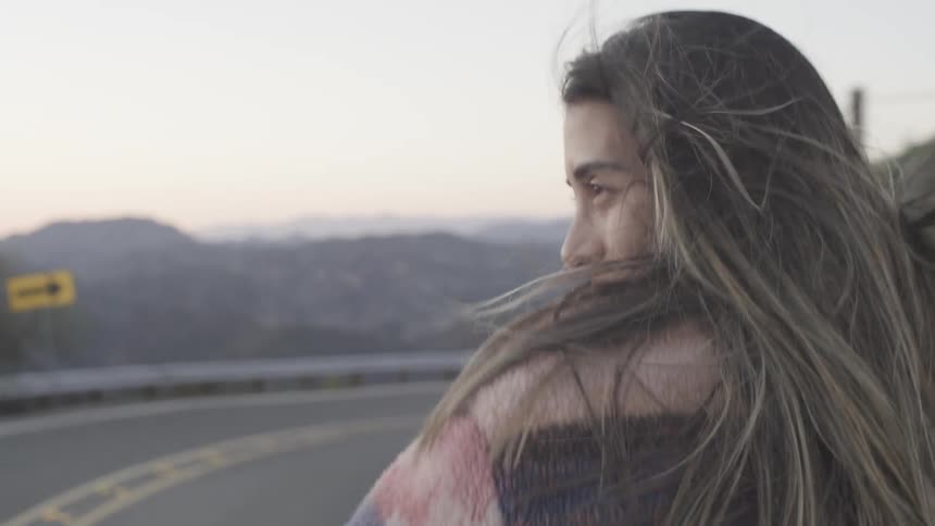 A woman leans out of a car sunroof smiling as wind blows through her hair on a mountain road at dusk