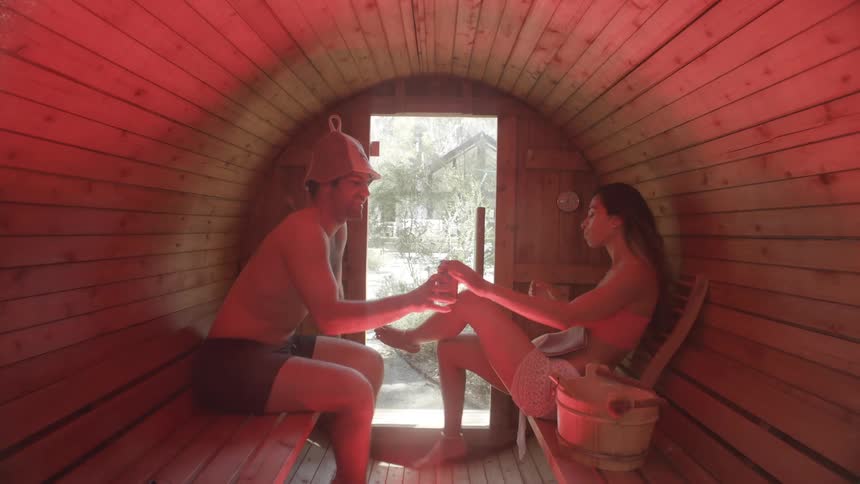 A man wearing a pointed hat and a woman in a light colored bikini sit inside a small wooden sauna while sharing a drink from a silver can