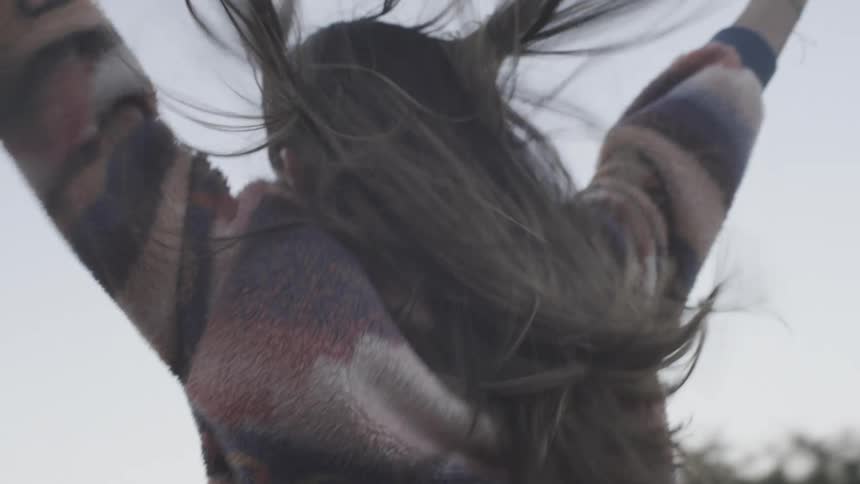 A woman with long hair and a patterned sweater spins joyfully outdoors under a pale skyas she turns toward the camera her hair catches the wind and she reveals a bright happy smile