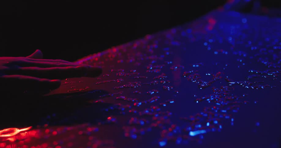 A close up view shows a hand as it touches a wet surface with red and blue light in a dark area while it rains