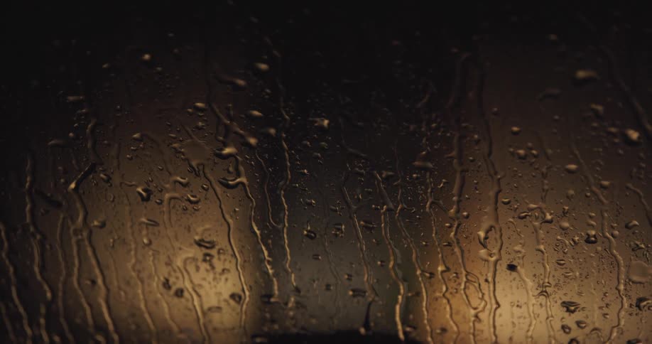 Rain drops slide down a glass surface with a golden light shining from behind in a dark setting
