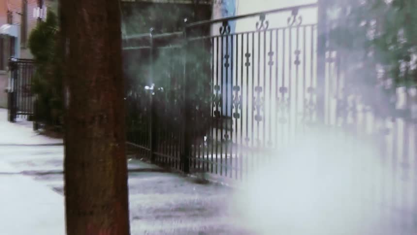 From a shaky perspective steam billows from a city sidewalk grate while two people holding an umbrella walk past a metal fence