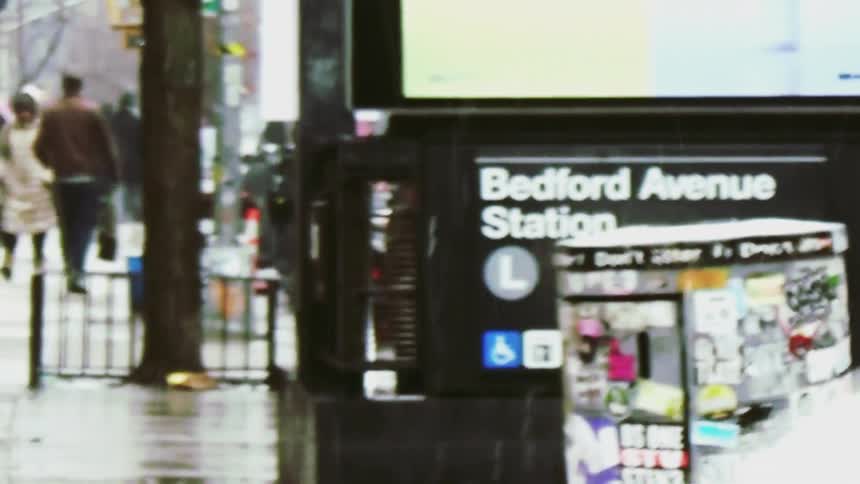 A view shows the entrance to the bedford avenue subway station in new york city while people walk on a wet sidewalk and cars drive past on a rainy day