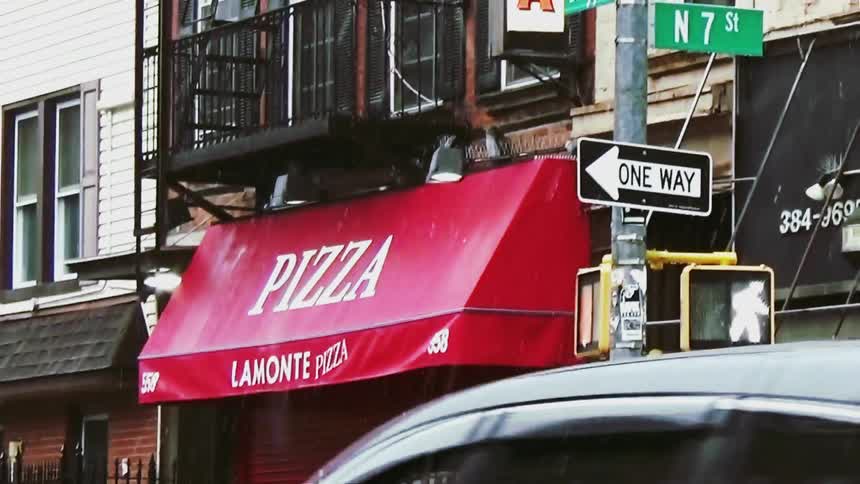 Rain falls over a city street corner featuring lamonte pizza located at the intersection of driggs avenue and north 7th street the scene captures a red storefront awning and a vertical pizza sign positioned next to a black fire escape on a brick buildin
