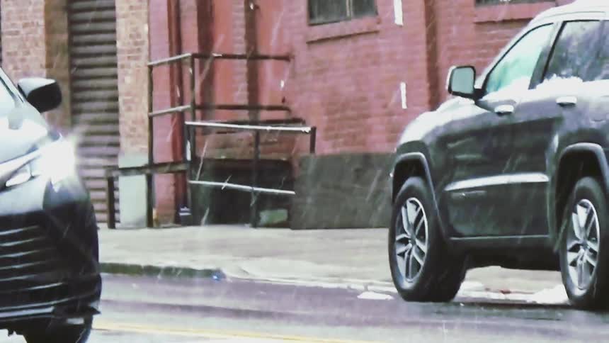 A woman wearing a grey coat and a white hard hat walks across a city street while carrying a white bagas she crosses a black toyota and a dark suv are parked or idling nearby while light snow falls in the background