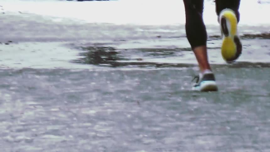 A view from behind shows the legs and feet of a person wearing black leggings and white sneakers running away from the camera on a wet and icy sidewalk