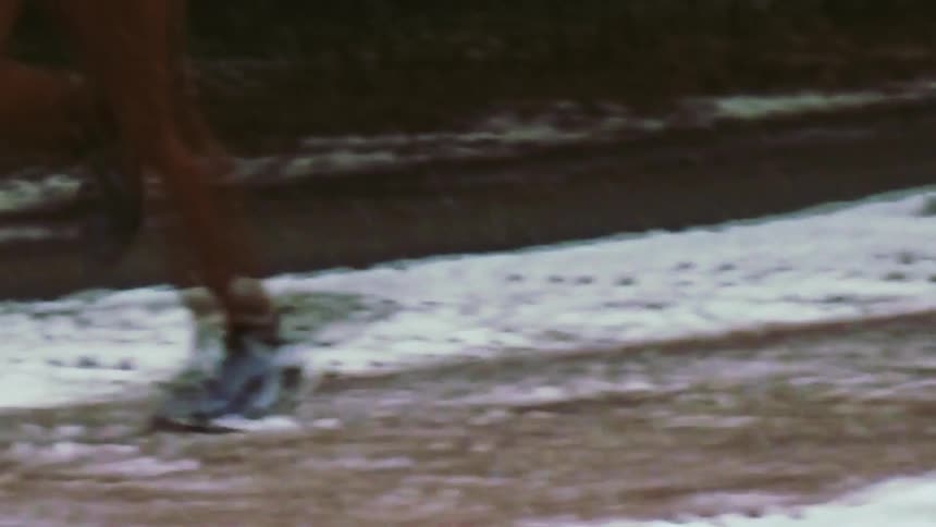 A view from the side shows the legs of a person wearing brown pants and blue sneakers running across a snowy and icy ground during winter
