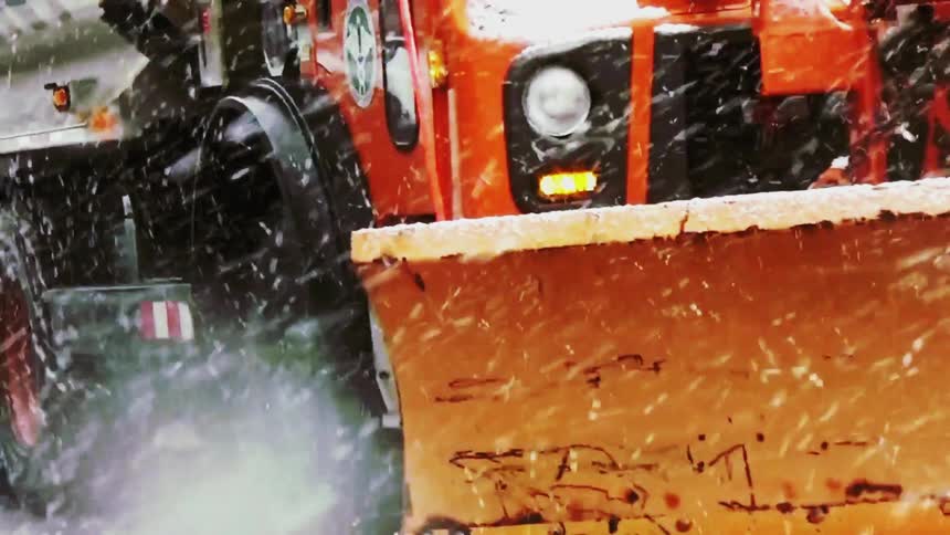 An orange snowplow truck drives through a heavy snowstorm clearing the road with its large front bladethe camera focuses on the trucks wheels as they spin through the slush and falling snow