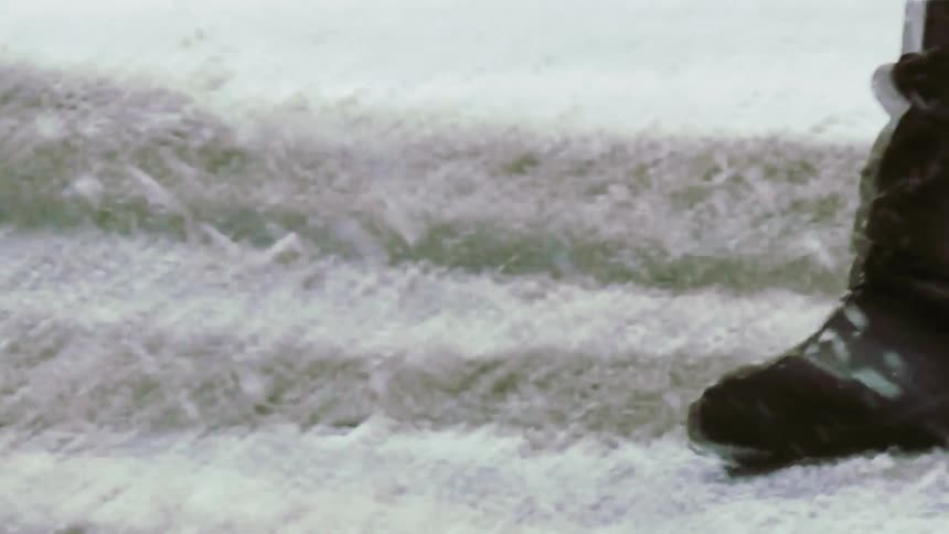 A close view of a person wearing black boots and blue jeans walking across a snow covered street during a winter storm