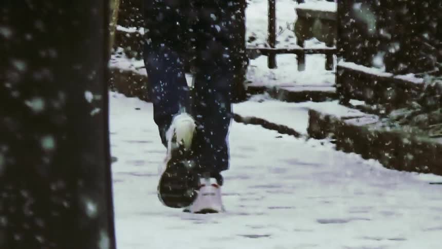 A view from behind shows the legs of a person wearing blue jeans and white sneakers walking away from the camera on a sidewalk covered in snow during a winter storm