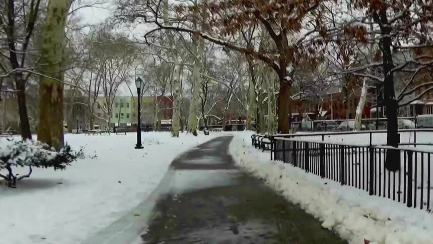 A view shows a paved path winding through a park covered in white snow with bare trees and a black metal fence on a cold winter day