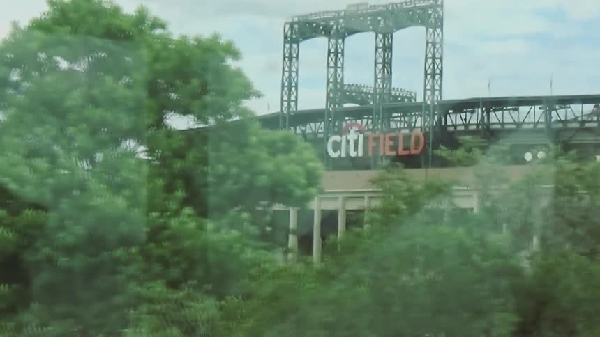 A view through a window shows a large stadium with the words citi field on the side while trees and a parking lot filled with cars pass by under a cloudy sky
